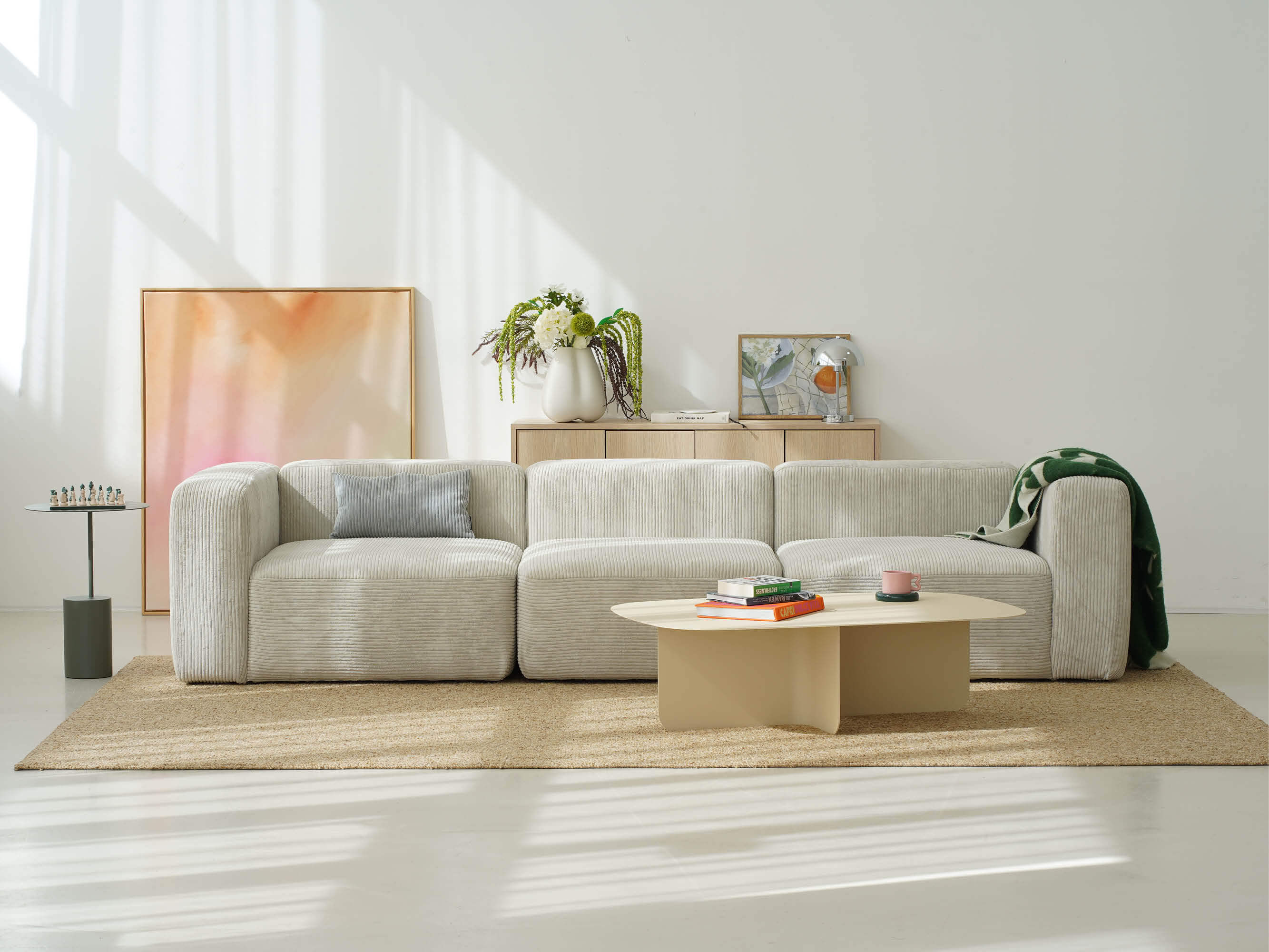 Modulares Cord-Sofa in hellem Beige als Viersitzer aus drei Elementen, aufgestellt in einem lichtdurchfluteten Wohnzimmer auf einem Juteteppich, davor ein heller Couchtisch mit Büchern, im Hintergrund ein Sideboard aus Eiche und ein abstraktes Kunstwerk in Rosatönen.