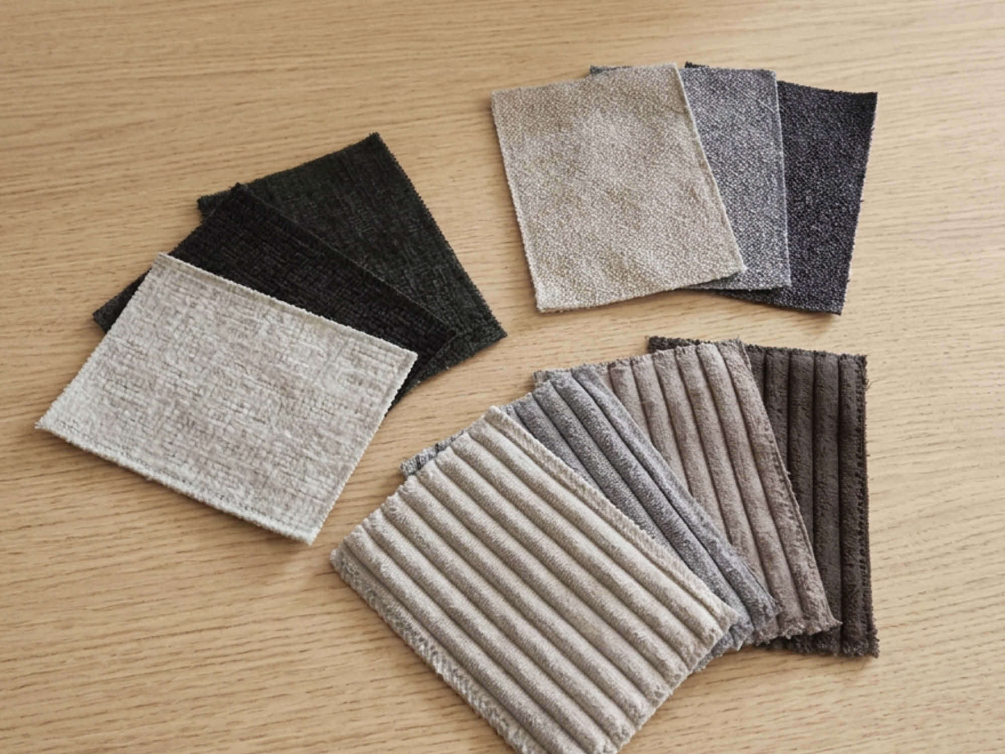 Stoffmuster-Auswahl auf einer hellen Holzfläche – mehrere Gruppen von Bezugsstoffen in Cord, Webstoff und Chenille in den Farbtönen Beige, Grau, Dunkelgrün und Braun.