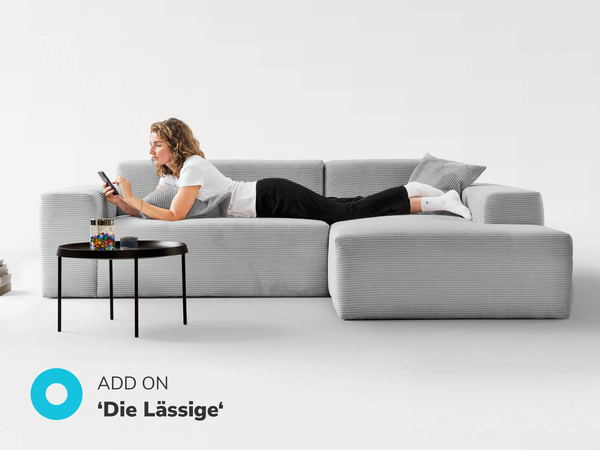 Cordsofa Ecksofa in grau, Ottomane rechts, Frau liegt auf dem Sofa und tippt auf ein Handy, Add on 'Die Lassige' 