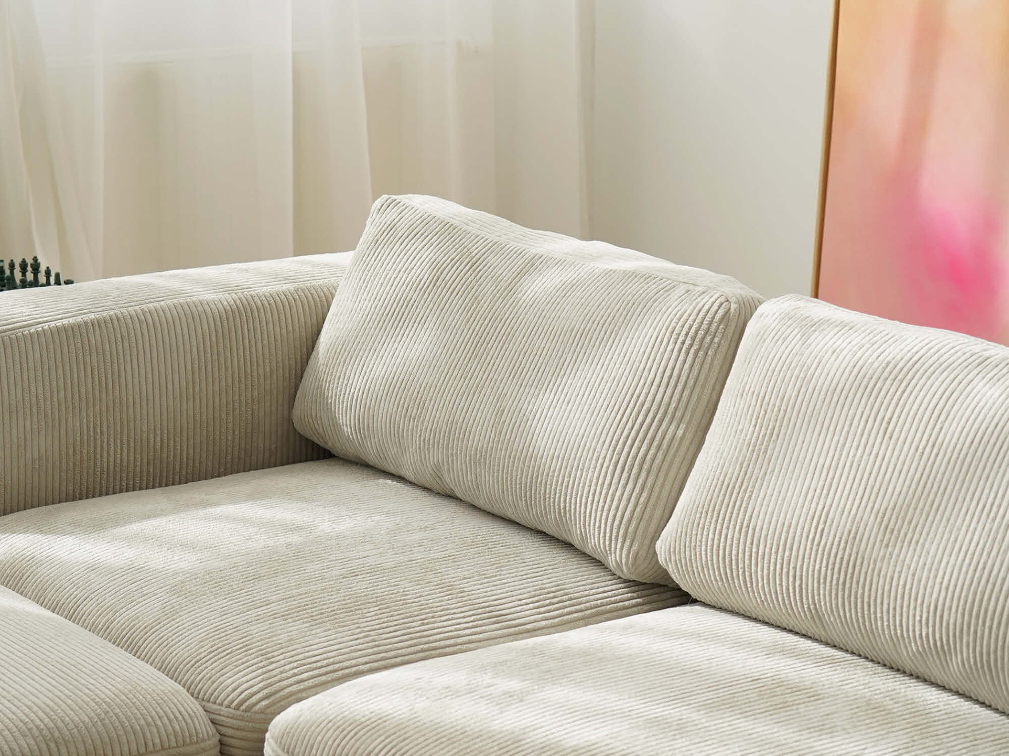 Detailansicht der Rückenkissen des Cord-Sofas in hellem Beige mit deutlich sichtbarer Cord-Rillenstruktur, im Hintergrund zartes Tageslicht und ein rosafarbenes Kunstwerk.
