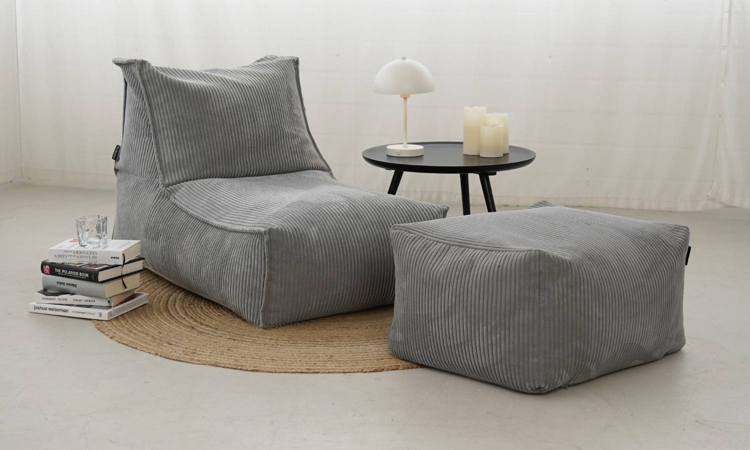 Sitzsack in Cord hellgrau mit Pouf 'Der Gelassene' auf einem runden braunen Teppich, Couchtisch un Bücherstapel daneben 