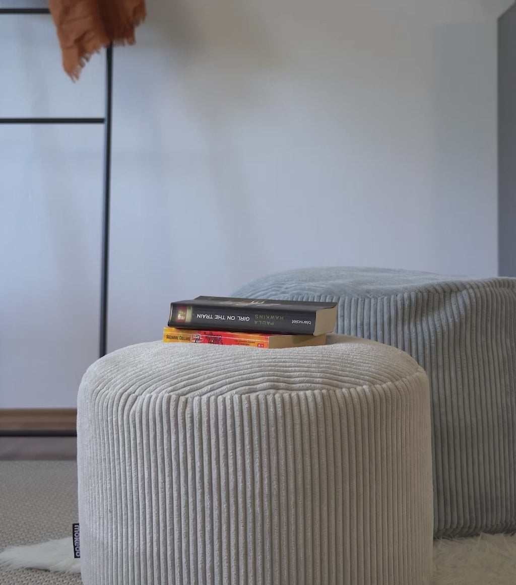 Produktvideo Pouf Sitzhocker aus Cord. Zwei Poufs in beige und grau. Bücher liegen auf dem Pouf, die Beine werden auf den Pouf gelegt und am Ende wird eine Tasse Tee auf den Pouf abgestellt. 